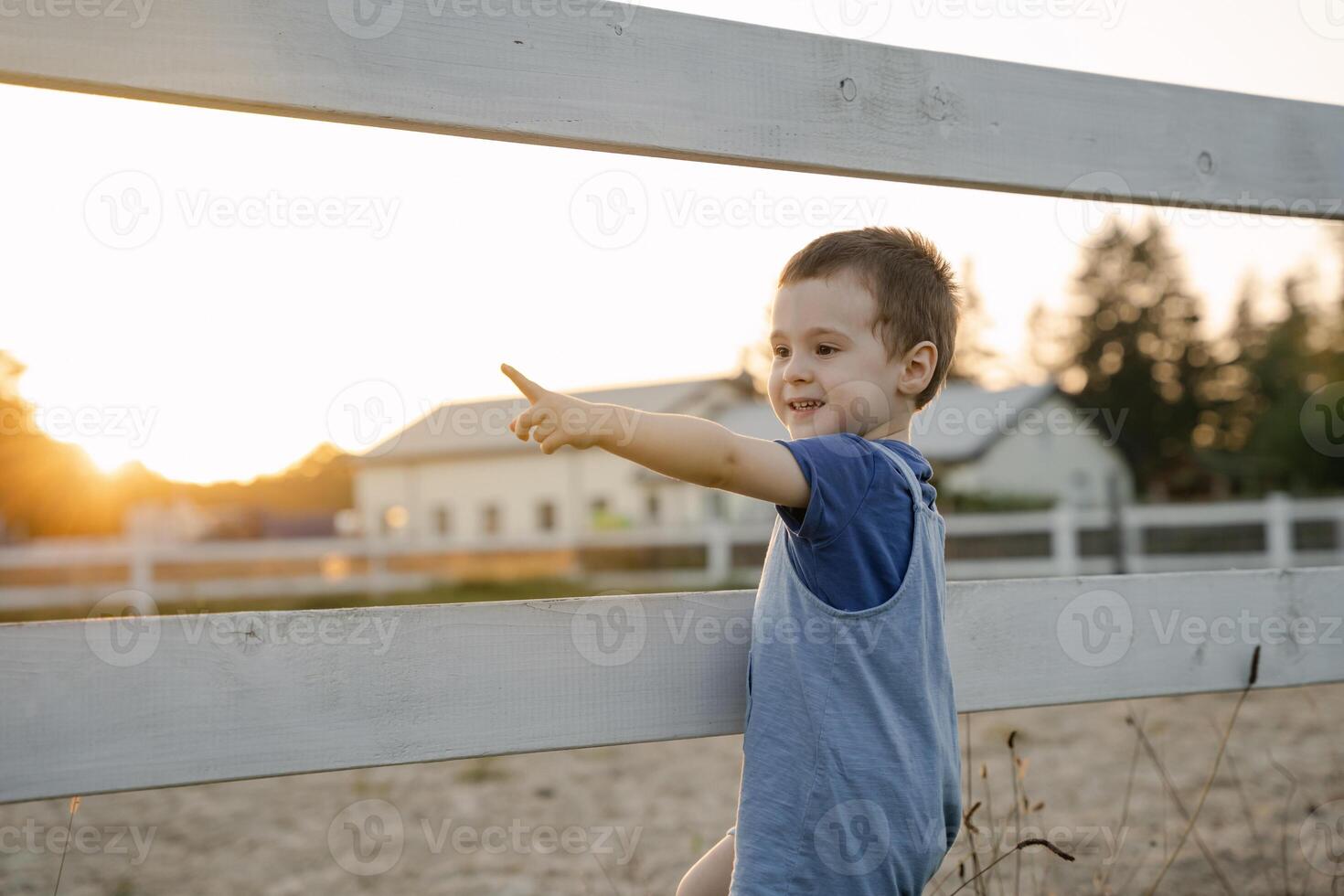 Happy toddler boy two or three years old stands near a fence and points with his finger on a sunny summer day. Summer sunset light. Horse farm photo