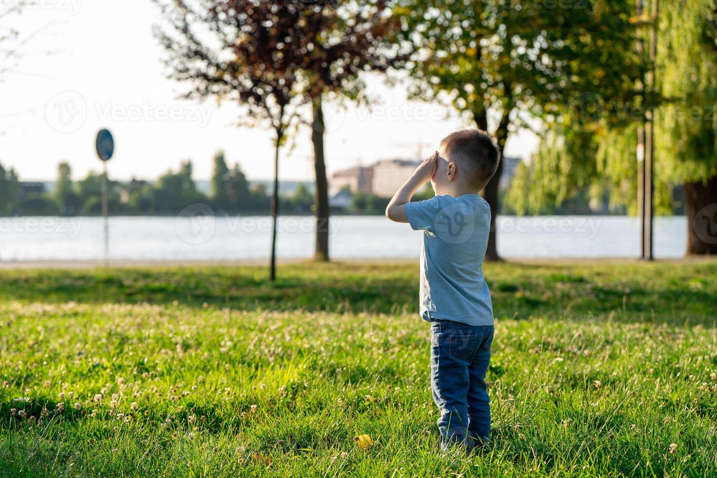 A little toddler boy of three and a half years old is standing in a summer park near a lake with his eyes closed. Childhood and nature. photo