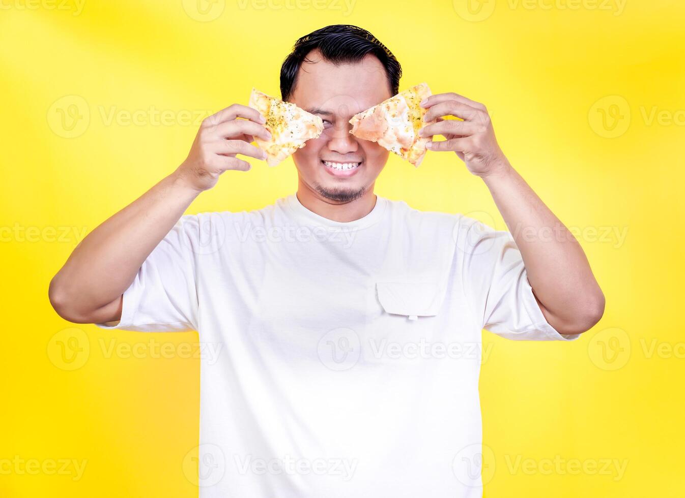 A cheerful asian man playfully holds pizza slices in front of his face against a vibrant yellow backdrop. international foods day concept photo
