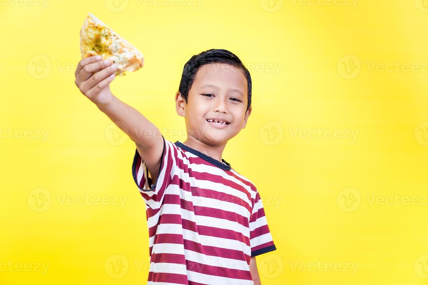 A cheerful asian boy holds up a slice of pizza against a bright yellow background, spreading joy. international foods day concept photo