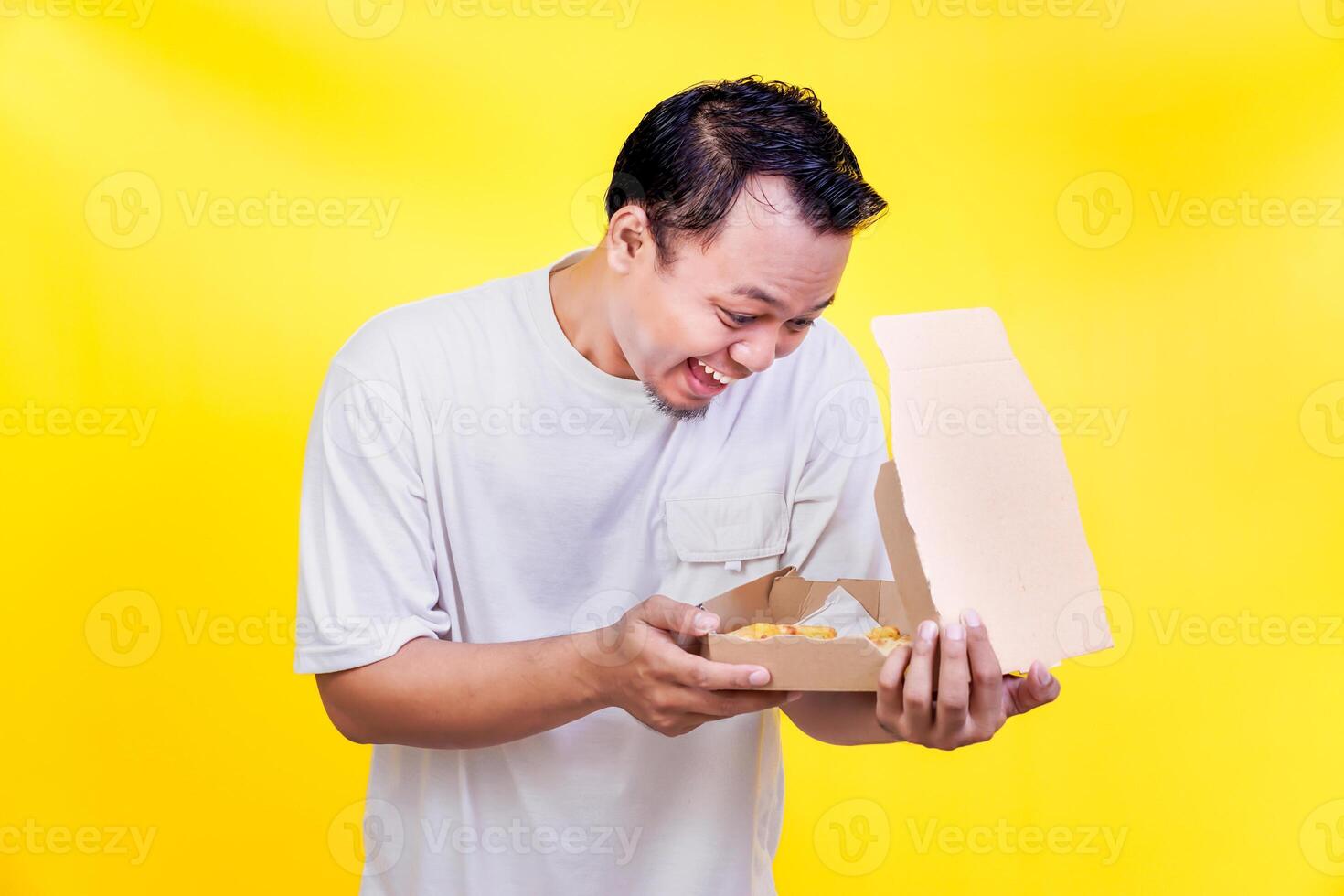 A happy asian man joyfully opens a takeout box, revealing delicious food against a bright yellow backdrop. international foods day concept photo