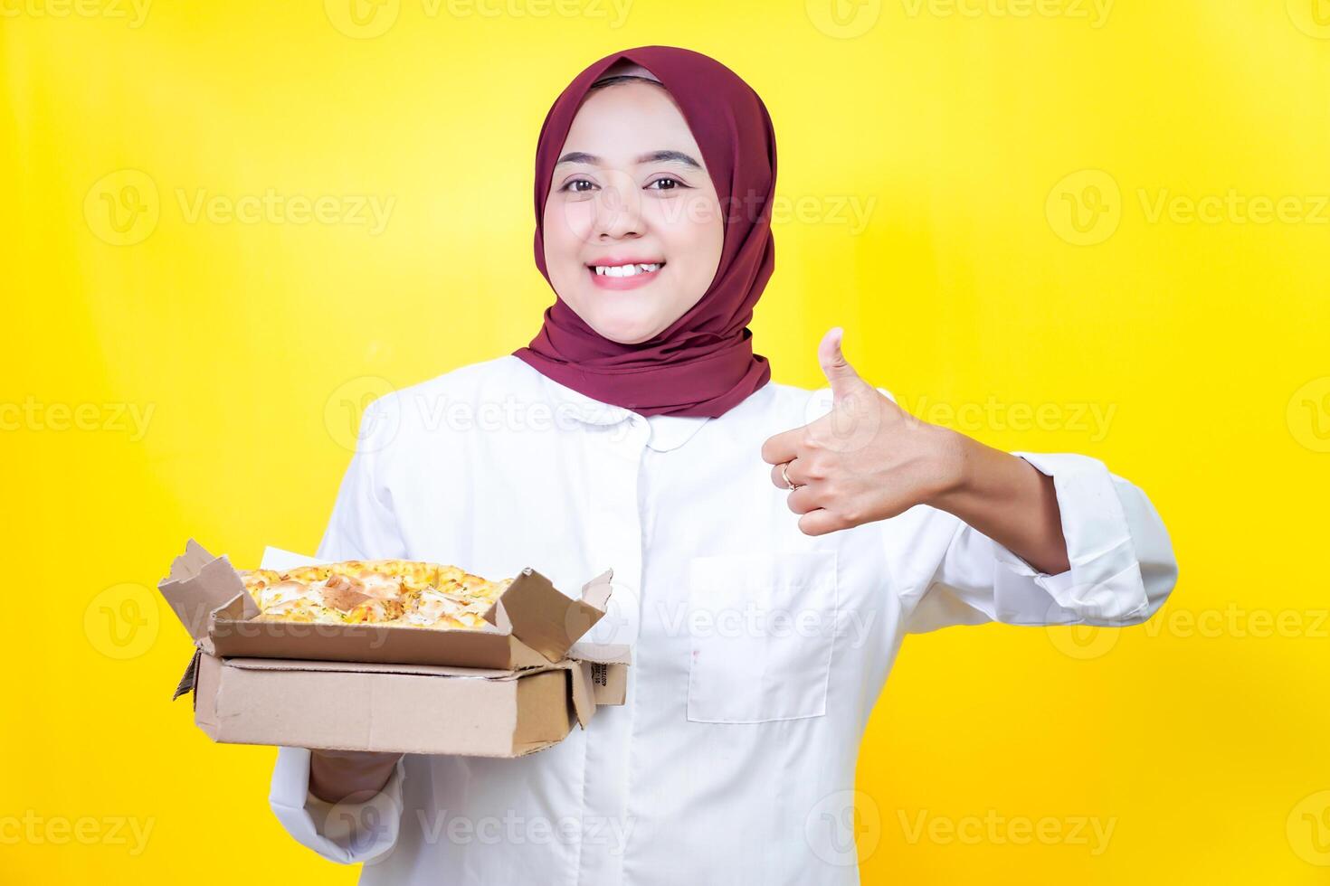 A smiling asian woman in a hijab holds a pizza box, giving a thumbs up against a yellow background photo