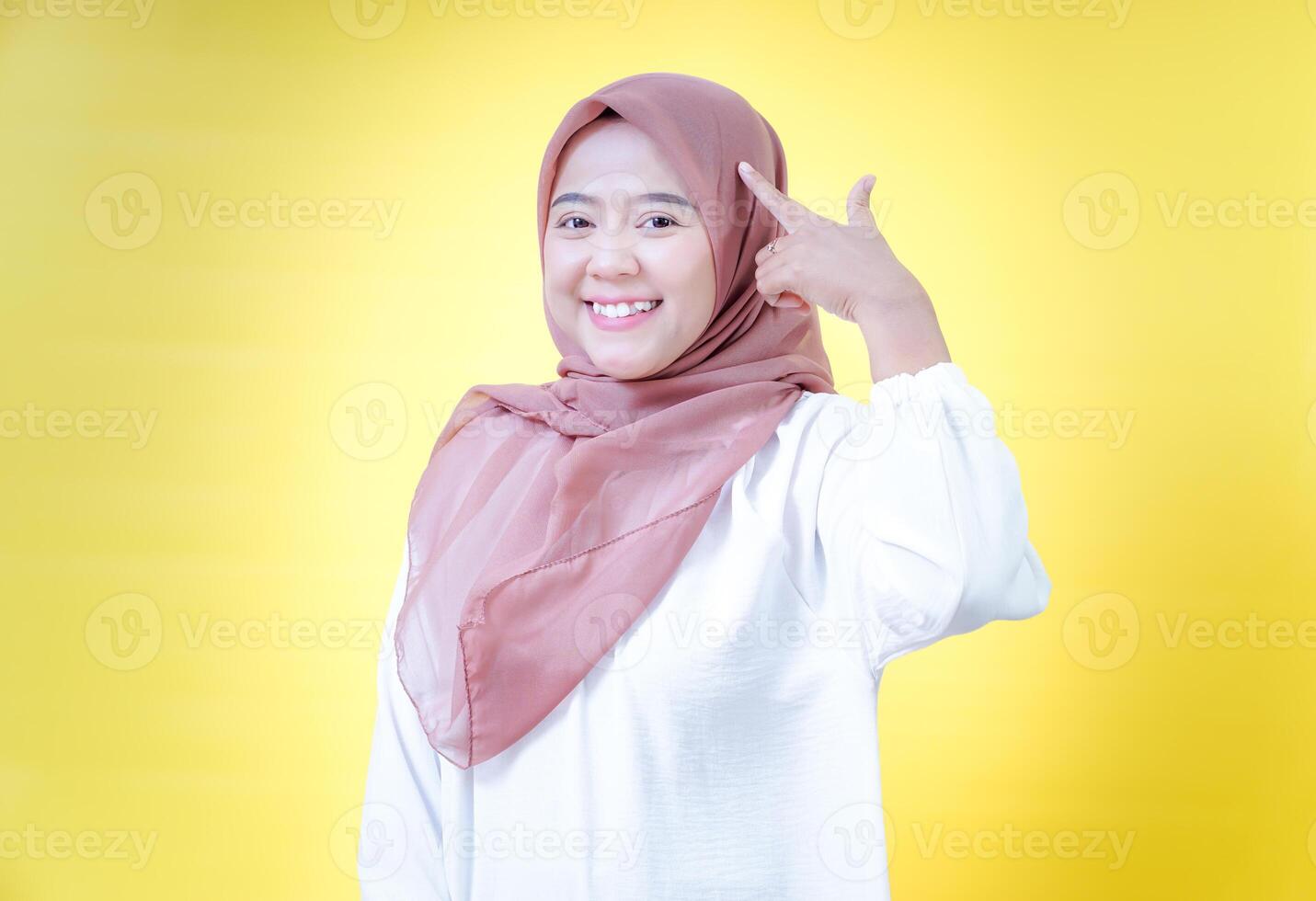 A joyful woman in a headscarf poses having idea by touching her head against a vibrant yellow background photo