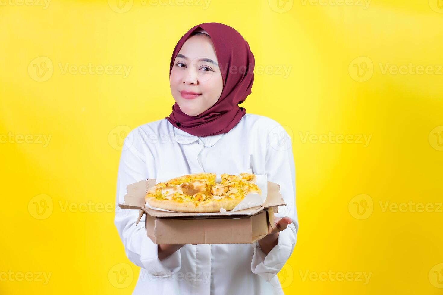 A smling asian woman in a hijab holds and showcasing a pizza box against a bright yellow backdrop photo