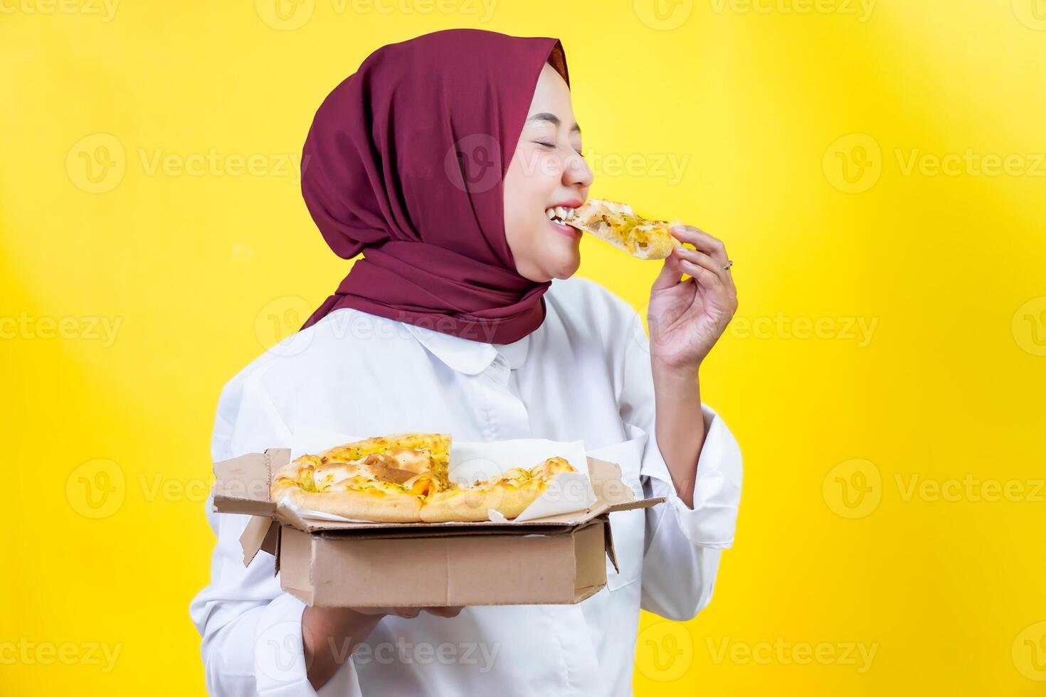 An asian woman bites a slice of pizza while holding a box of pizza in front of a bright yellow background. photo