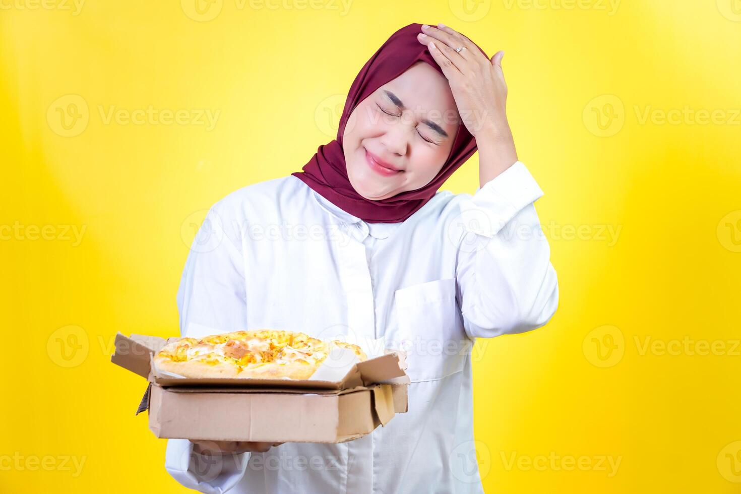 Happy asian woman in a hijab holding pizza with a playful expression against a bright yellow background photo