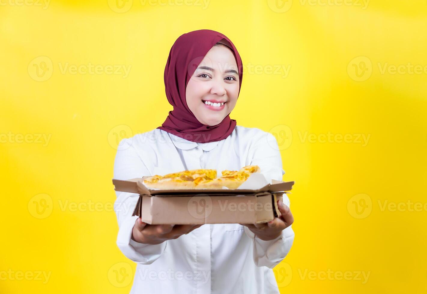 A beautiful asian woman in a hijab holds and showcasing a pizza box against a bright yellow backdrop photo
