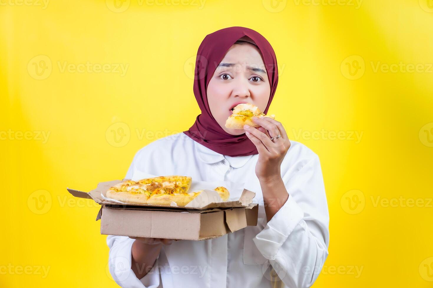 An asian woman in a hijab enjoys a slice of pizza while holding a box of pizza in front of a bright yellow background. photo