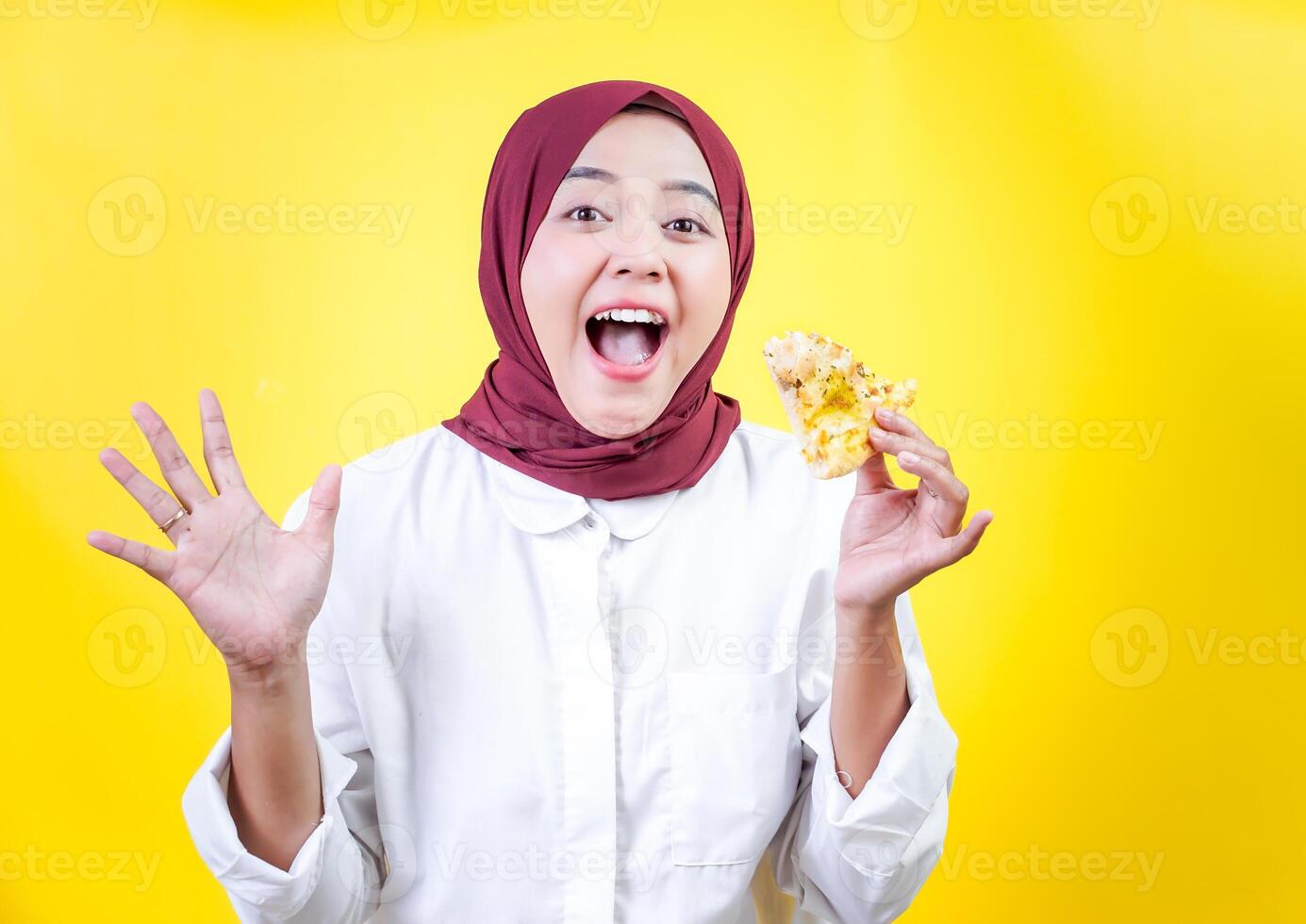 A joyful asian woman in a white outfit and hijab holds a tasty snack against a yellow background photo