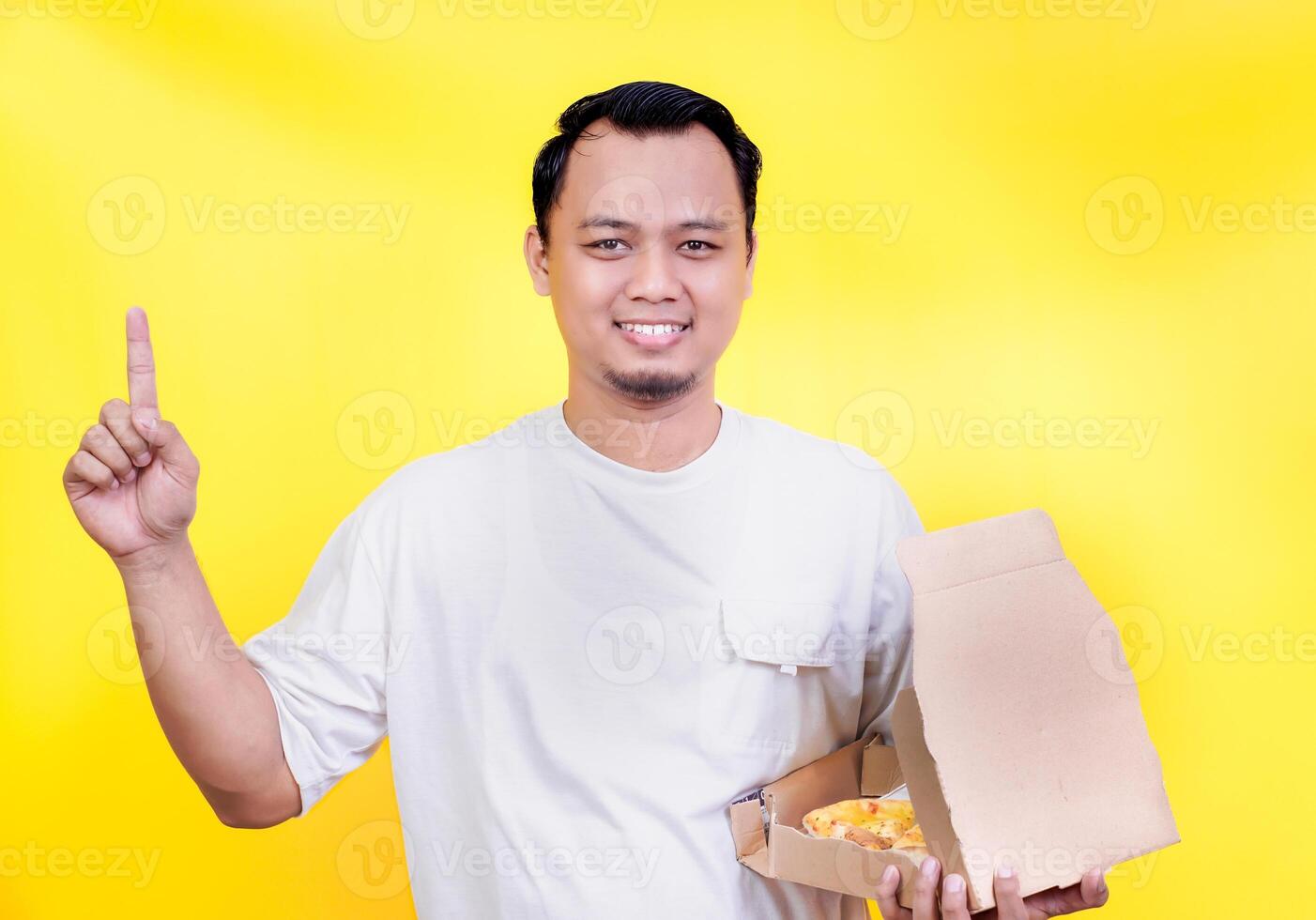 A smiling asian man in a white shirt holds a pizza box, pointing up against a vibrant yellow background. international foods day concept photo