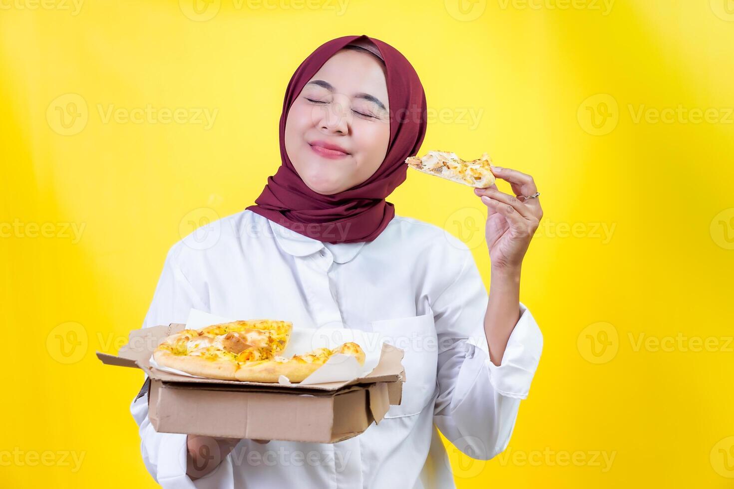 An asian woman njoying a delicious slice of pizza with a joyful expression against a yellow background photo