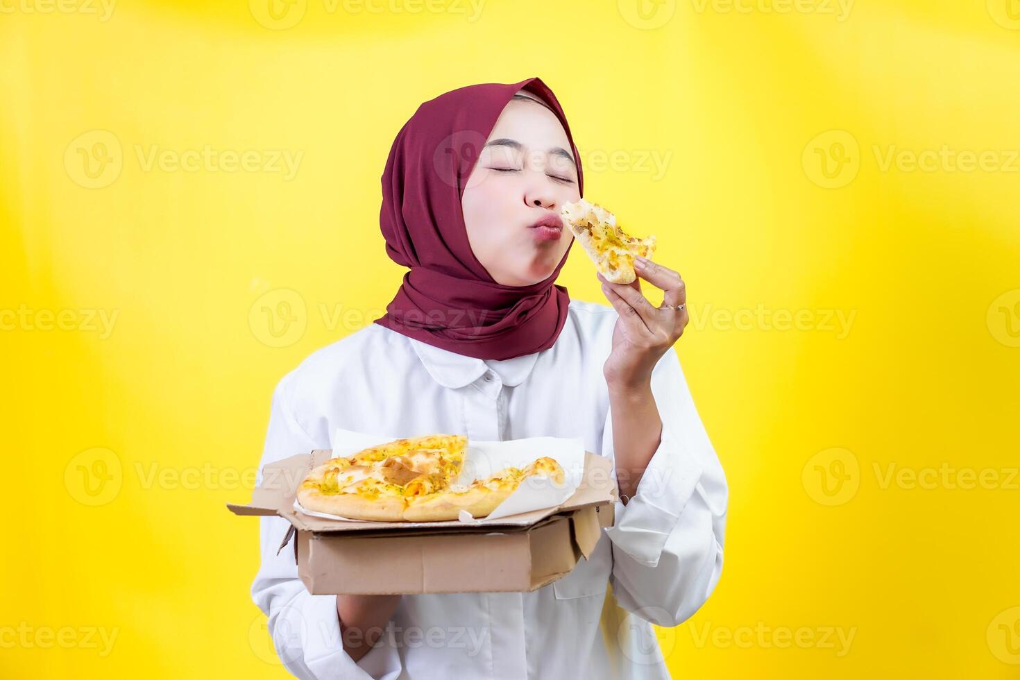 An asian joyful woman in a hijab enjoys pizza isolared on a vibrant yellow background photo