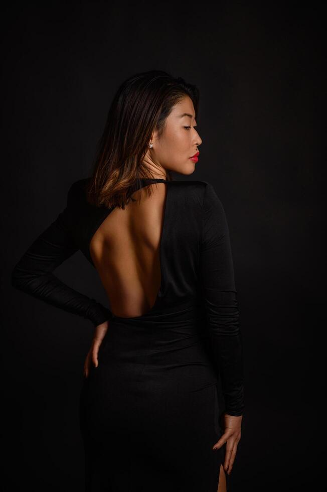An Asian woman in a black dress exudes elegance as the dark studio setting amplifies her presence. photo