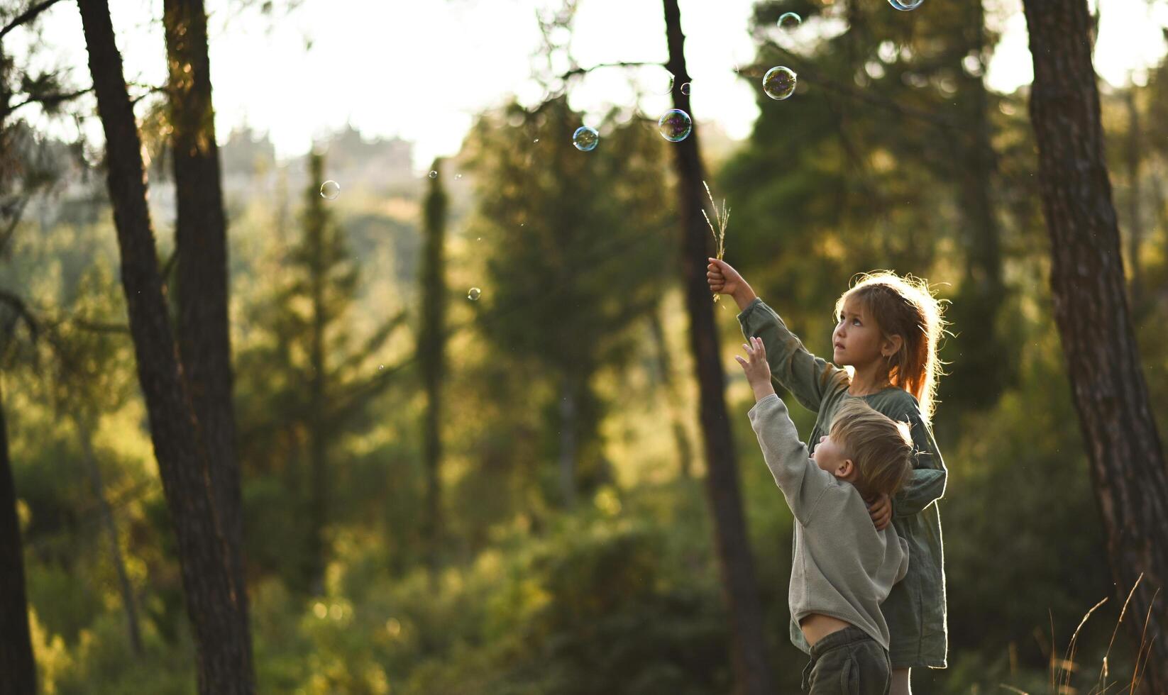 In the heart of a magical woodland, a boy and girl stand surrounded by shimmering bubbles, their eyes filled with wonder. photo