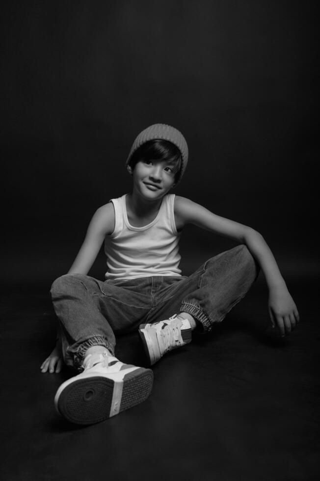Against a deep black background, an Asian boy in a white tank top and jeans stands with a natural and effortless stance, highlighting his youthful energy. photo