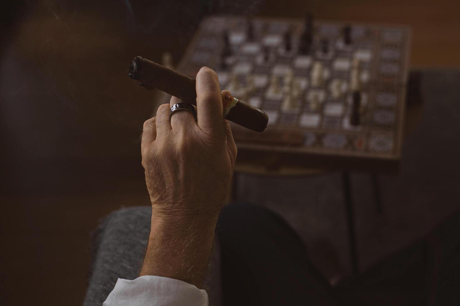 A steady hand holds a smoldering cigar, wisps of smoke curling above the chessboard, where a game of strategy unfolds in the dim light. photo