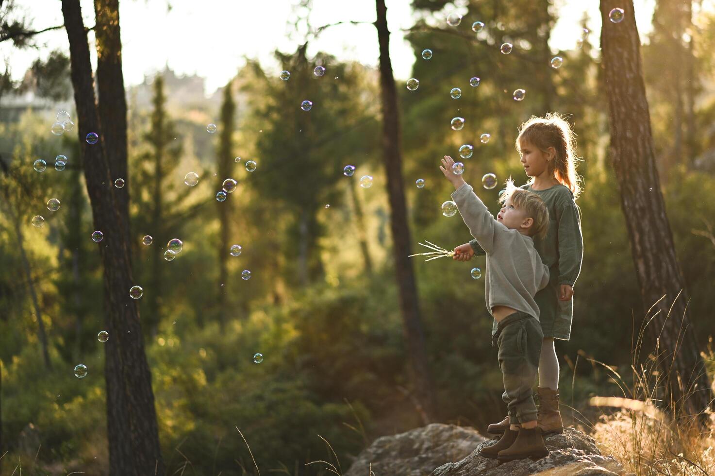 Among towering trees and soft moss, a boy and girl sit quietly, watching as shimmering bubbles drift like tiny fairy lights around them. photo