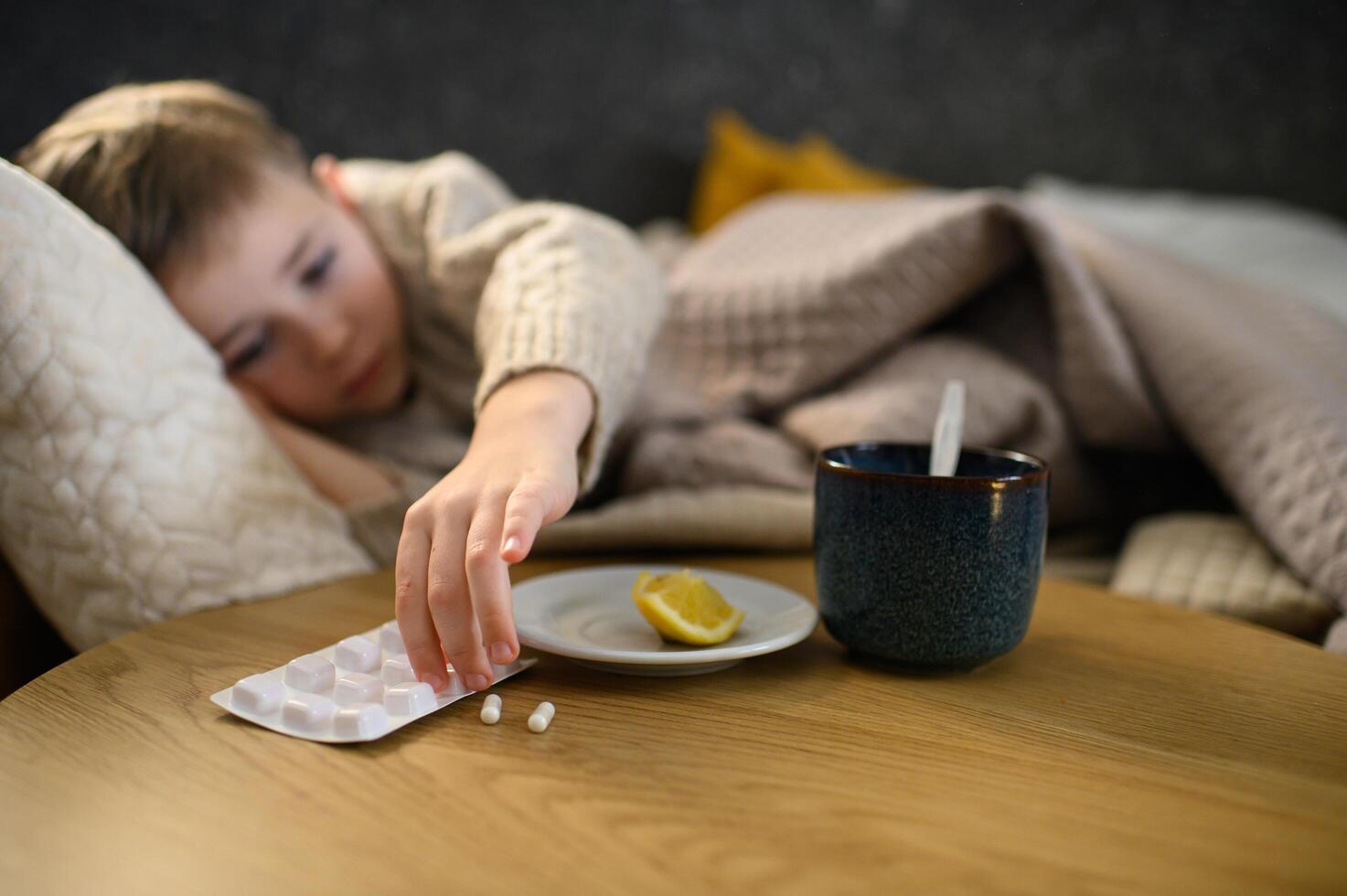 Looking exhausted, the boy lies in bed, his eyes heavy. A hot cup of tea with lemon and some pills sit nearby, offering comfort and relief. photo