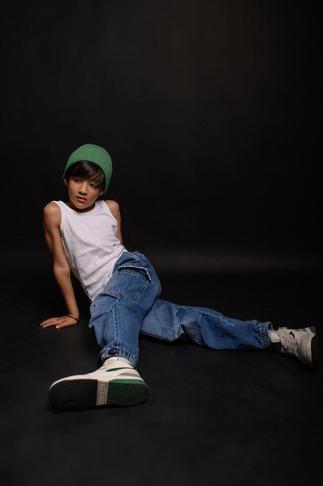 The boy, dressed in casual jeans and a white tank top, poses in a studio setting with a dark backdrop. His gaze is calm and focused. photo
