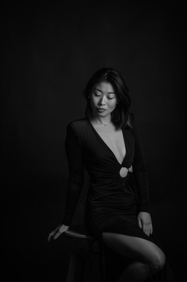 The minimalist lighting highlights the Asian womans beauty in a black dress, her pose commanding attention against the deep, dark backdrop. photo