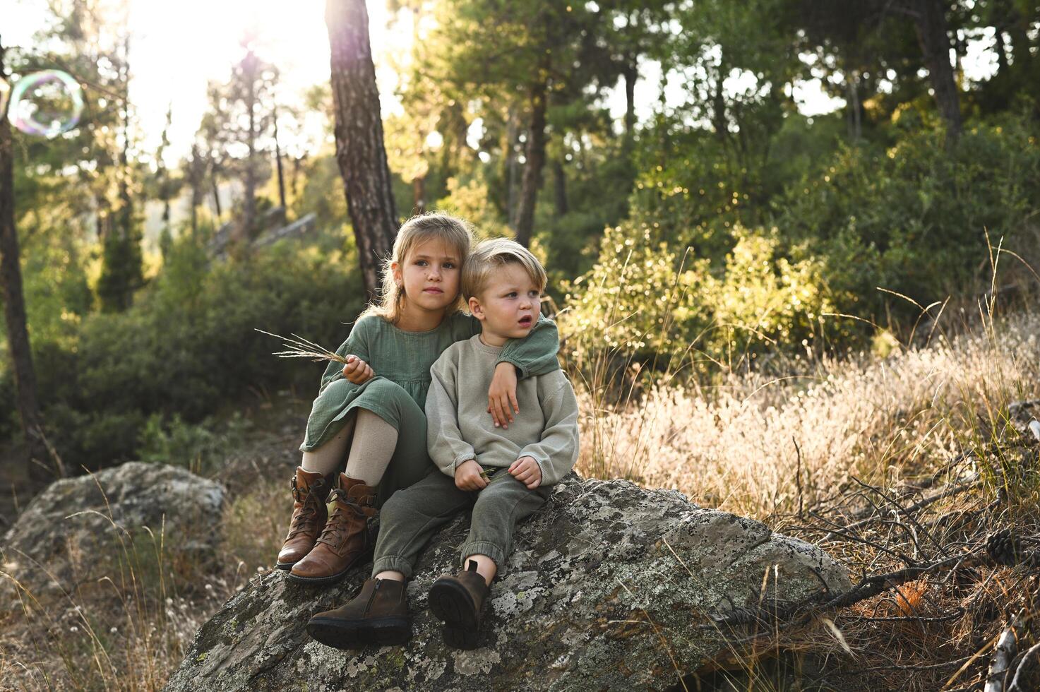 In a magical woodland, two children rest on a rock, watching delicate bubbles drift through the air like tiny, glowing orbs. photo