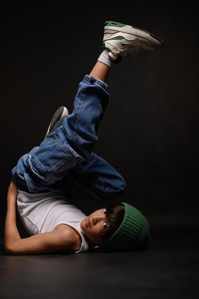 In a minimalist studio setting, an Asian boy in casual jeans and a white tank top poses confidently against a dark backdrop. photo