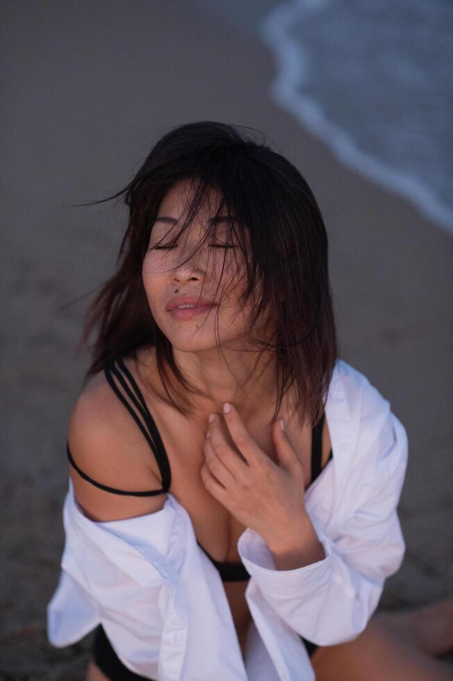 A beautiful Asian woman stands on the beach, wearing a black swimsuit and a white shirt, the ocean breeze gently lifting the fabric. photo