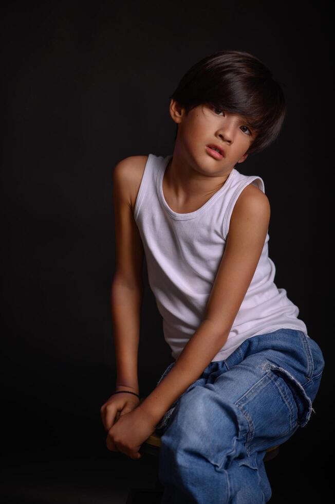 A young Asian boy stands against a black studio background, wearing a white tank top and jeans, his expression calm and composed. photo