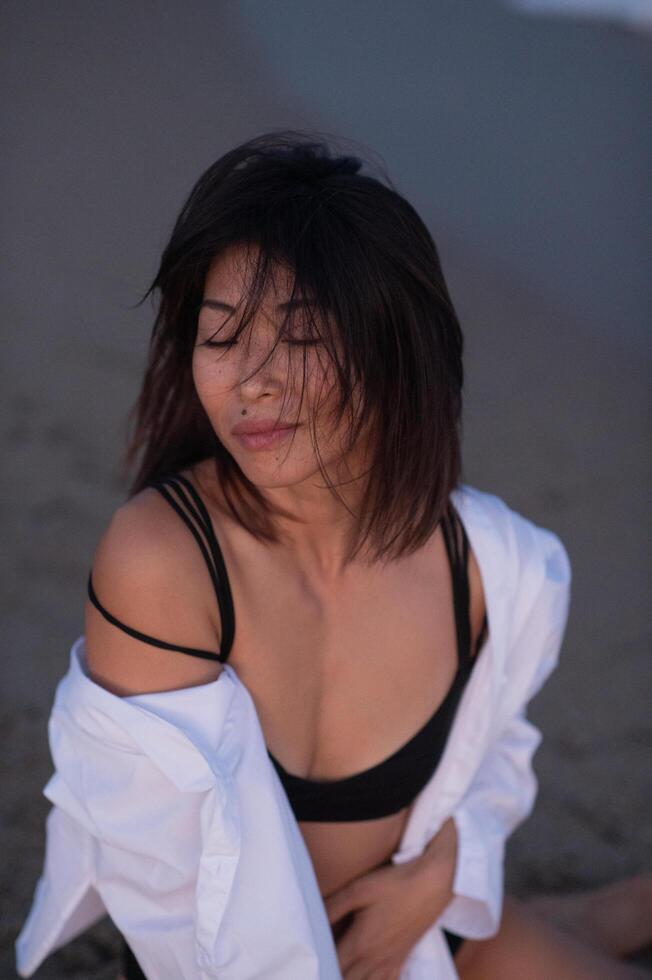 The breeze plays with her white shirt as an Asian woman in a sleek black swimsuit strolls along the beach, her steps light and graceful. photo