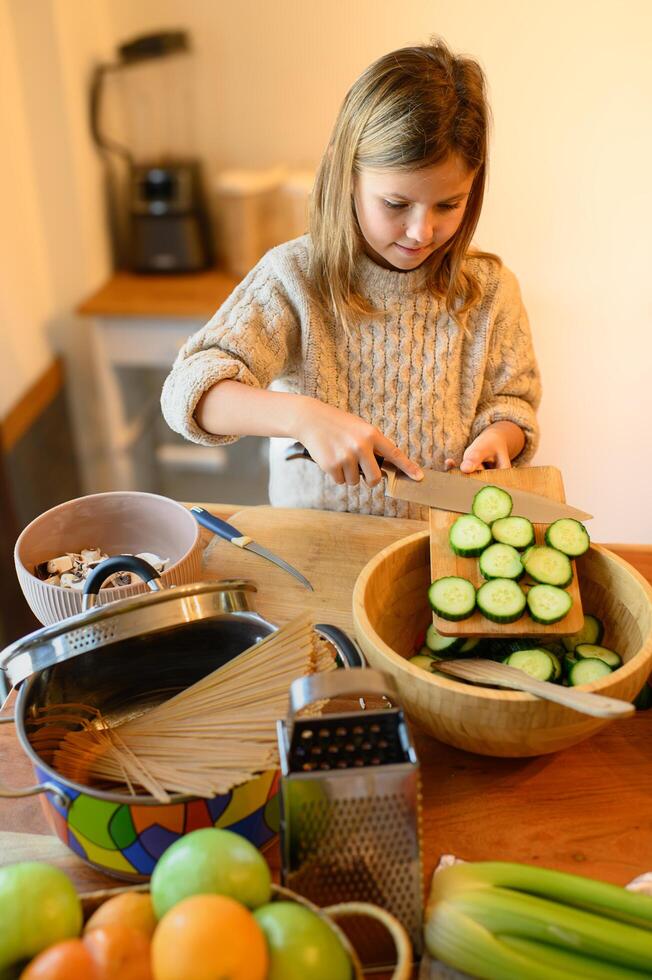 The girl carefully slices a fresh cucumber on a wooden cutting board, her small hands gripping the knife with focus as she tries to make even pieces. photo