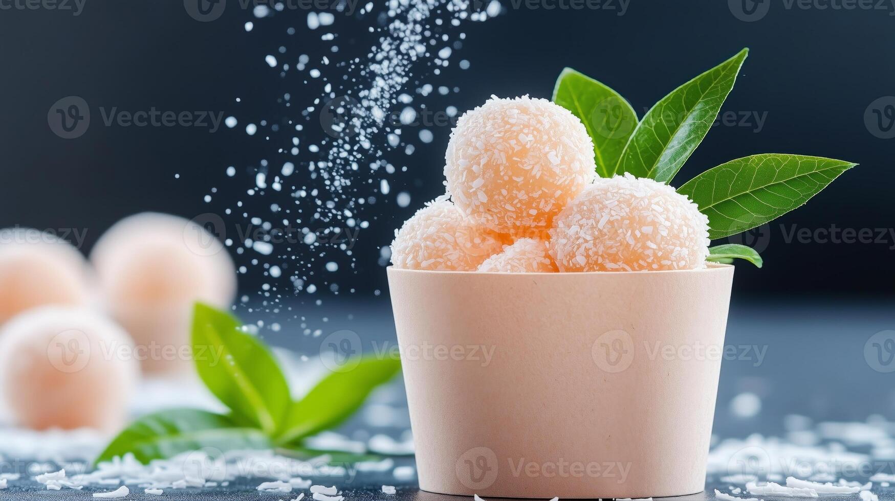 Close-up of coconut covered dessert balls in a paper cup with fresh green leaves, surrounded by falling sugar and a blurred dark background. photo