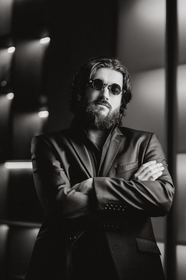 Portrait of handsome, young, confident bearded man adjusting in glasses, looking at camera while standing against dark studio background. Black and White photo