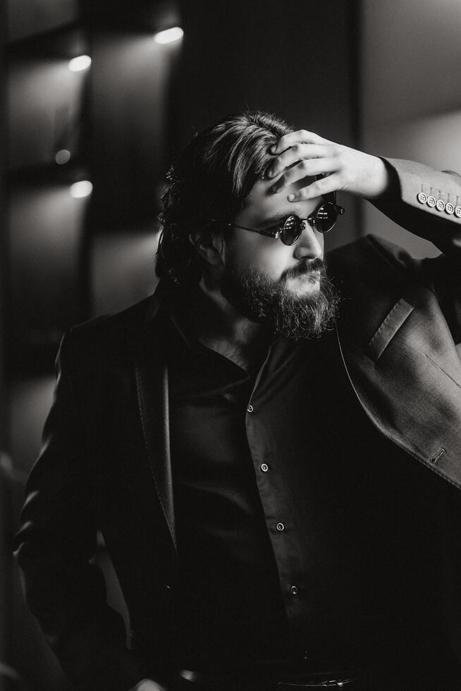 Side view Portrait of handsome, young, confident bearded man adjusting in glasses while standing against dark studio background. Black and White shot photo