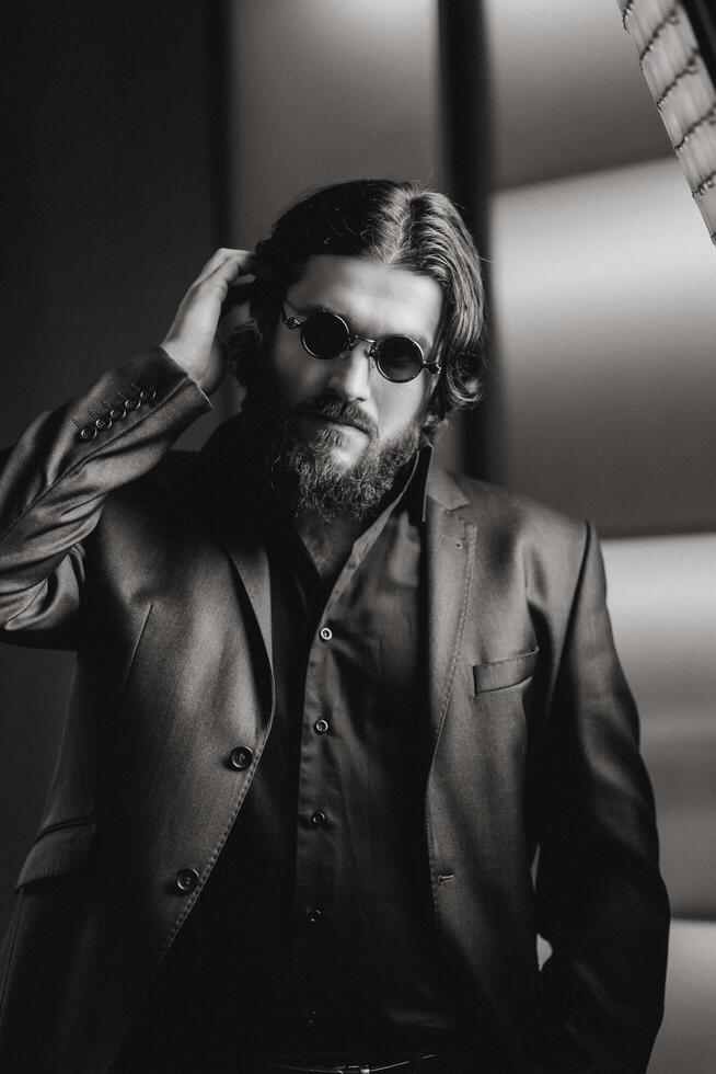 Side view Portrait of handsome, young, confident bearded man adjusting in glasses while standing against dark studio background. Cool fashion man in suit. Black and White shot photo
