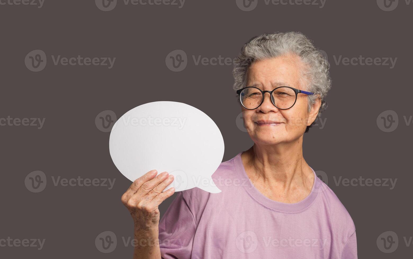Senior woman holding a blank speech bubble and looks at the camera with a smile while standing on a gray background. photo