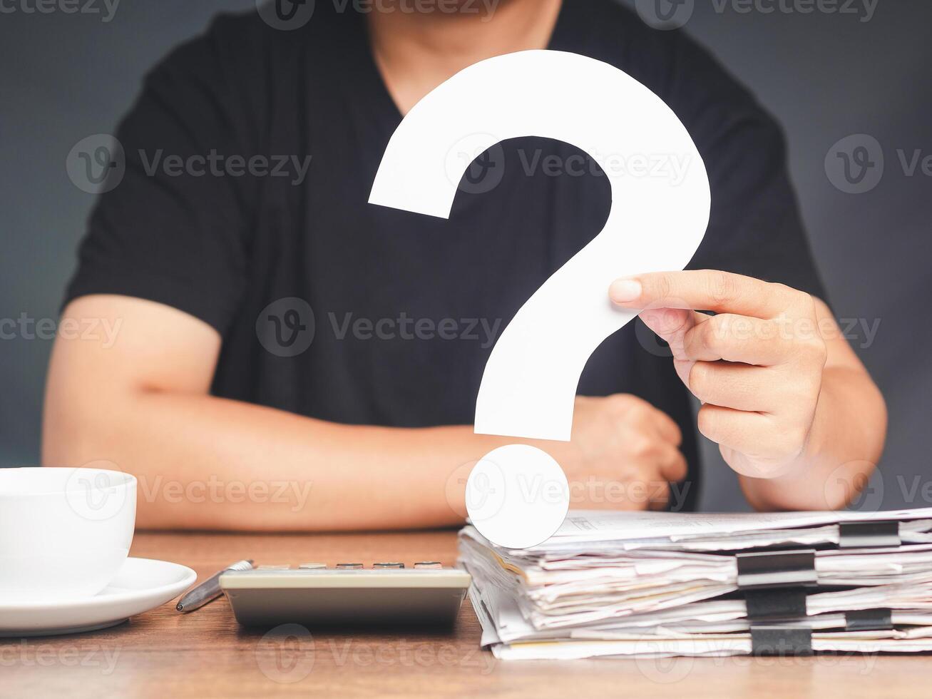 A young man in a black casual is holding a white question mark symbol while sitting at the table photo