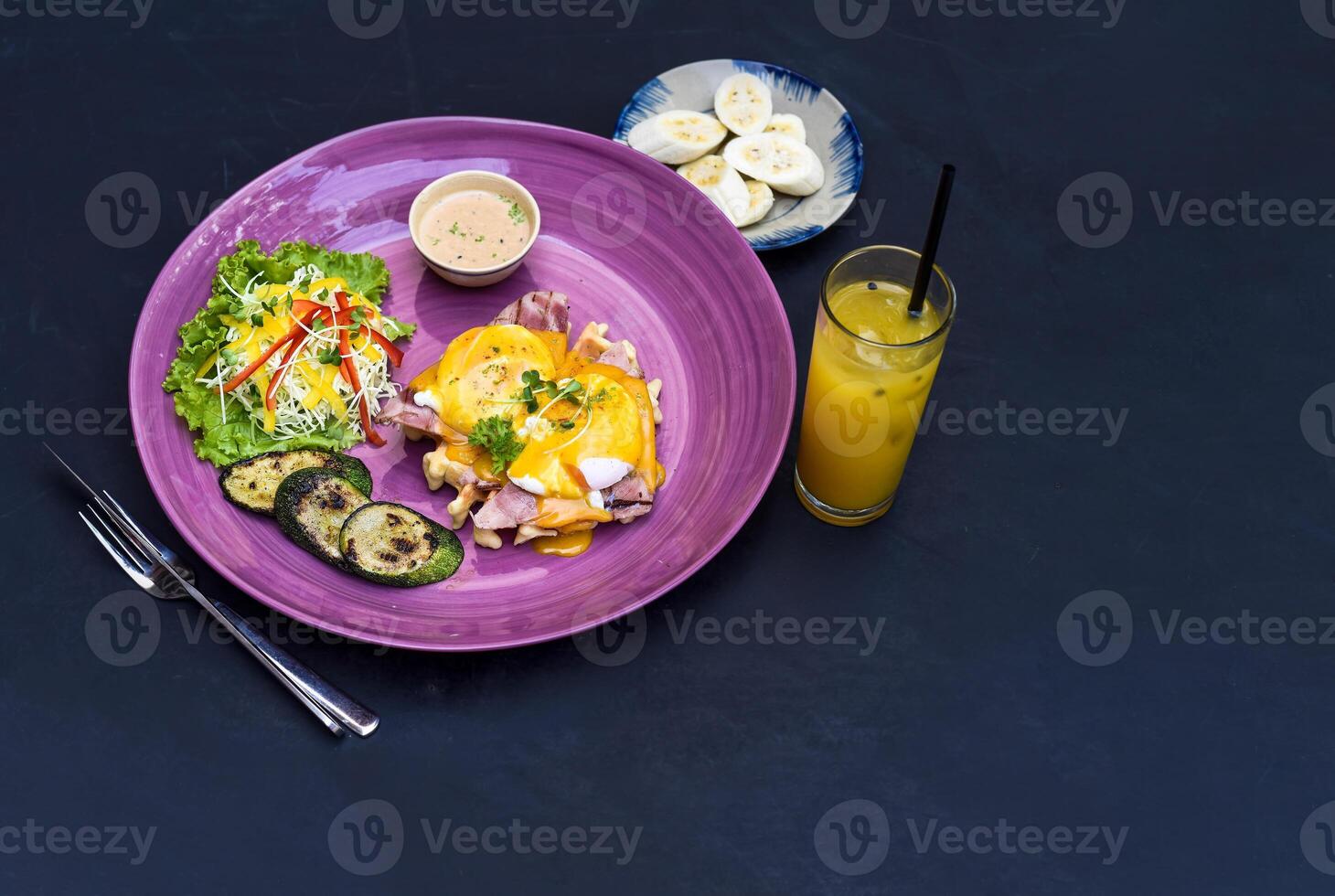 Eggs Benedict poached eggs on grilled ham with holland sauce on belgian waffle with grilled zucchini with plate of banana and cup of passion juice photo