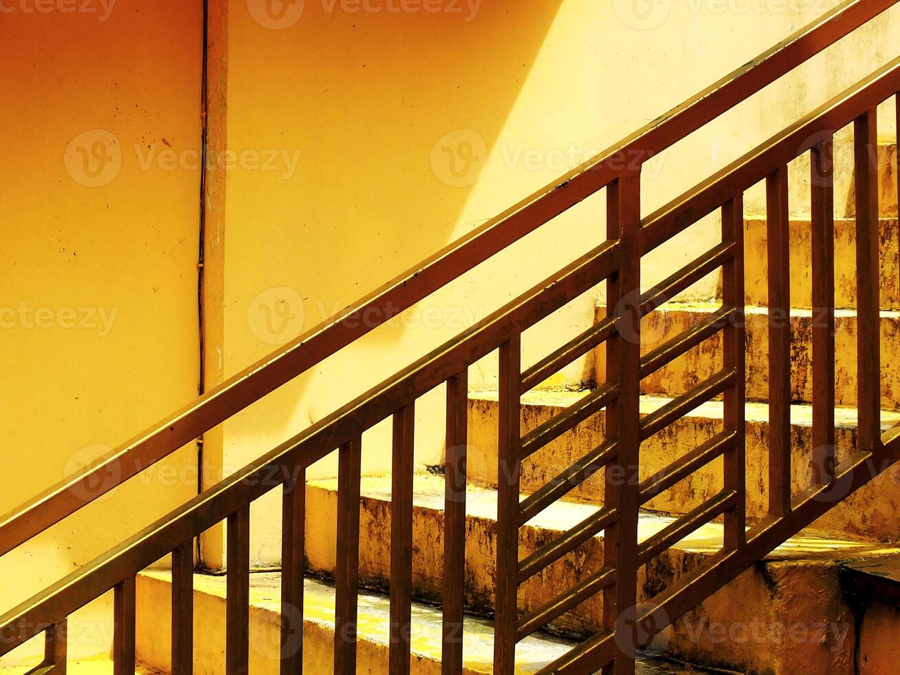View of a geometric abstract image of a staircase with a yellow wall in the background. The railing is made of metal and is painted gold photo