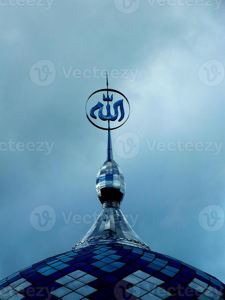 Blue and white mosque dome with an Arabic Calligraphy written Allah on top is on top of a building. The dome is located on a cloudy day. Islamic Ramadan Background. photo