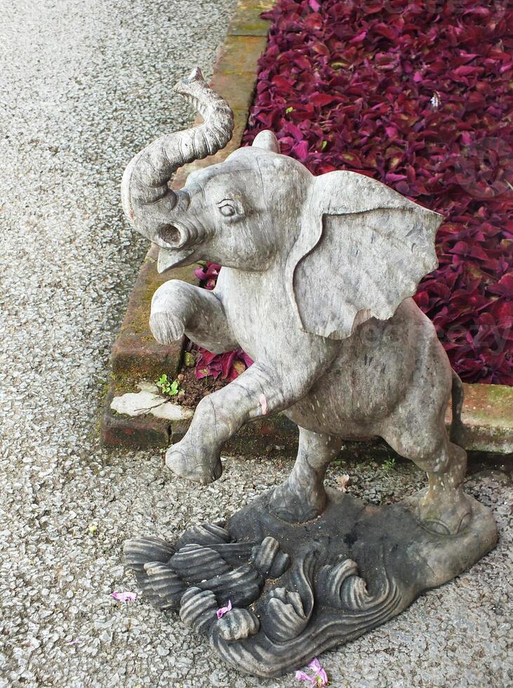 Grey elephant statue is standing on a stone path. The elephant is looking up. The statue is surrounded by red flowers, giving the scene a lively and colorful atmosphere photo