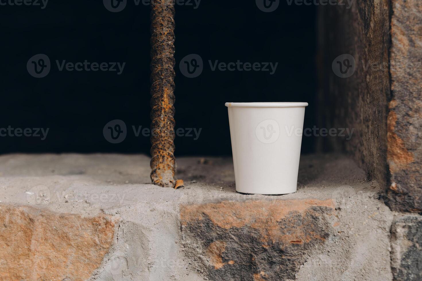 White disposable cup placed on a stone surface near a rusty rod in a dimly lit area photo