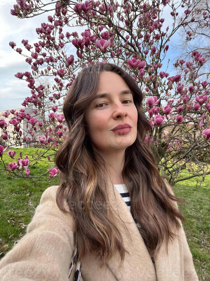Caucasian woman with air touch hair coloring taking a selfie near blooming magnolia tree in spring. photo