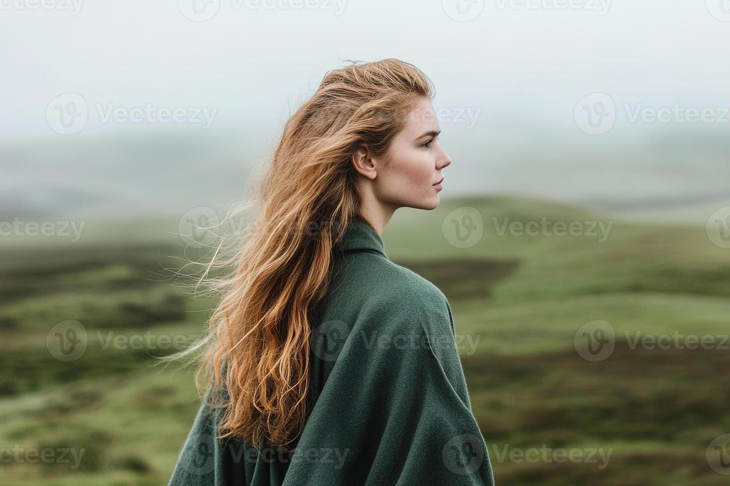 A woman stands amidst the lush Irish countryside, wearing a long green coat. She gazes thoughtfully at the rolling hills, with mist gently rising in the distance photo