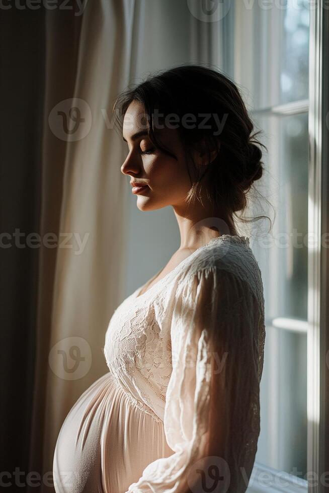 A pregnant woman stands by a window, softly illuminated by warm light. She gently cradles her growing belly, reflecting the beauty of motherhood in a serene moment photo