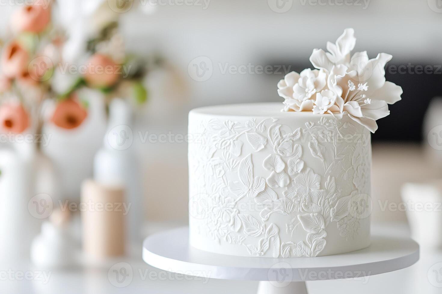 A smooth, white cake features delicate embossed floral details and is elegantly displayed on a stand. It complements a beautifully arranged setting of flowers, enhancing any celebration photo