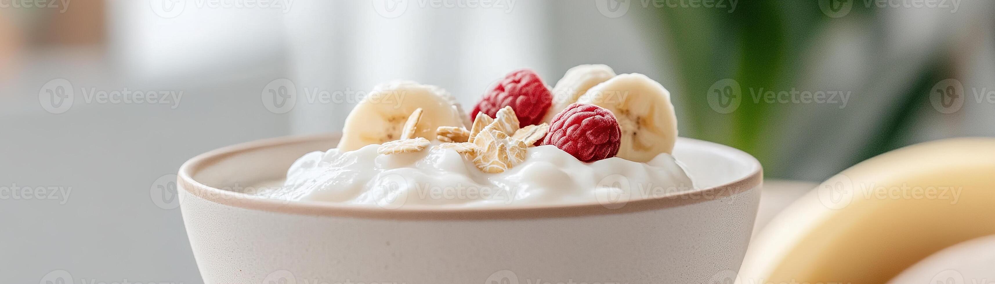 A softly illuminated bowl contains creamy yogurt adorned with banana slices and fresh raspberries. Natural light enhances the nourishing food, evoking a sense of gentle nutrition photo