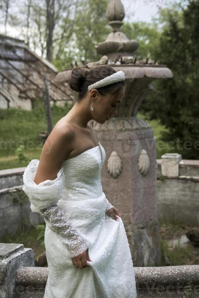 a beautiful girl in a white dress and a white tiara on her head walks against the background of an old 18th century building. the brunette has an elegant hairstyle, styled in a bun photo