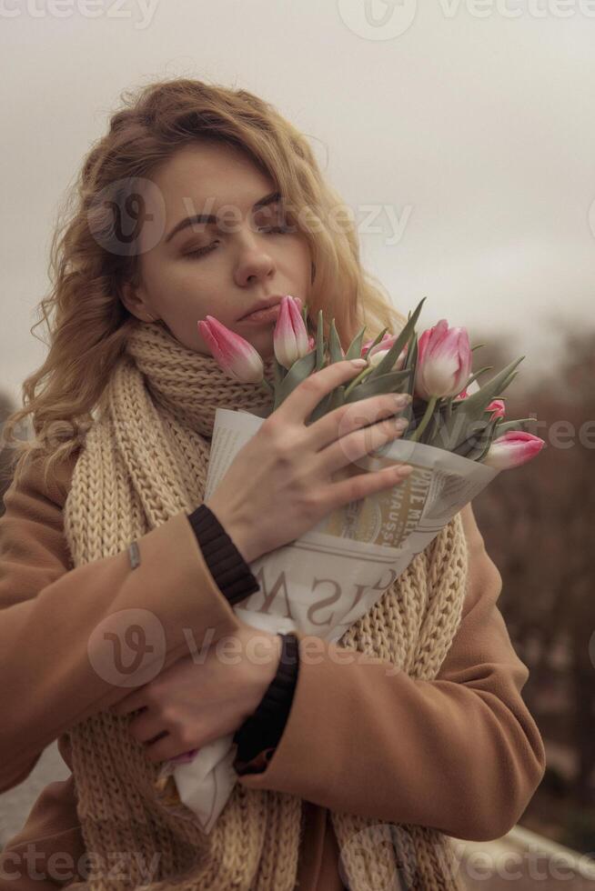 Close-up of a blonde with a bouquet of pink tulips. The bouquet is wrapped in craft paper. She is dreaming. Her hair is flying. She is wearing a beige classic coat and a warm scarf. photo