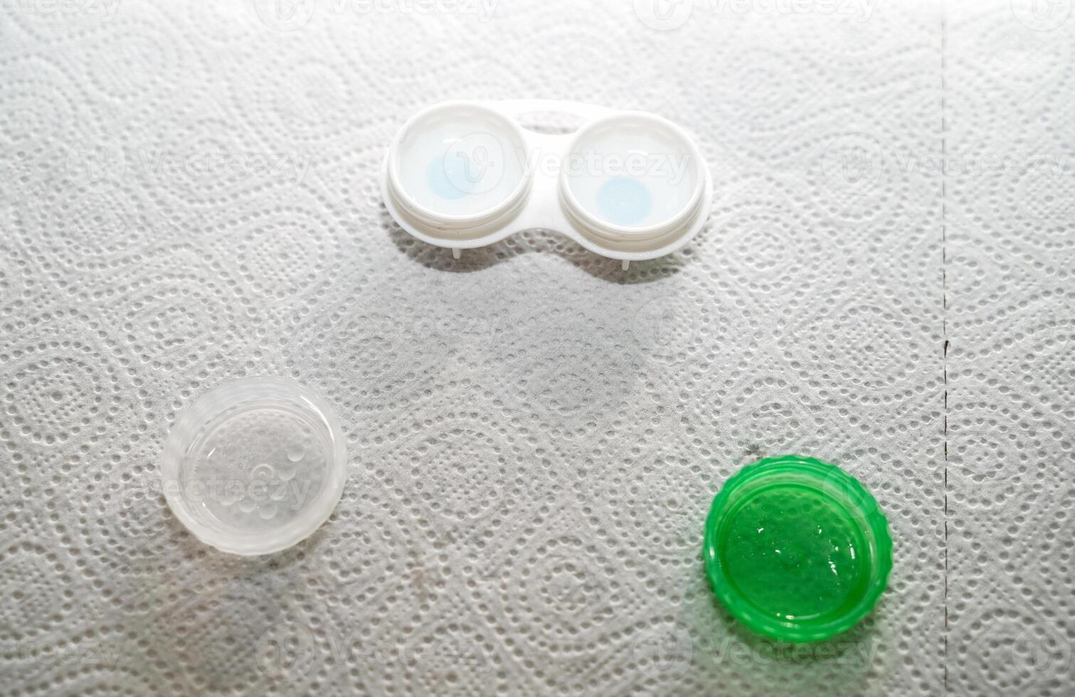 Container for storing lenses and contact lenses. The container is white with green, there are lenses in it. The table is covered with a paper towel for sterility at home. The container is open, you can see the lenses in it. photo
