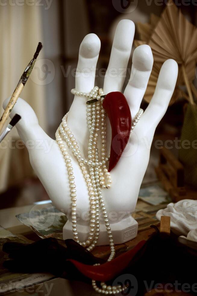 plaster hand holding pearl necklace and red hot pepper. close-up hand made of white plaster, paint brushes nearby. concept hot classic photo
